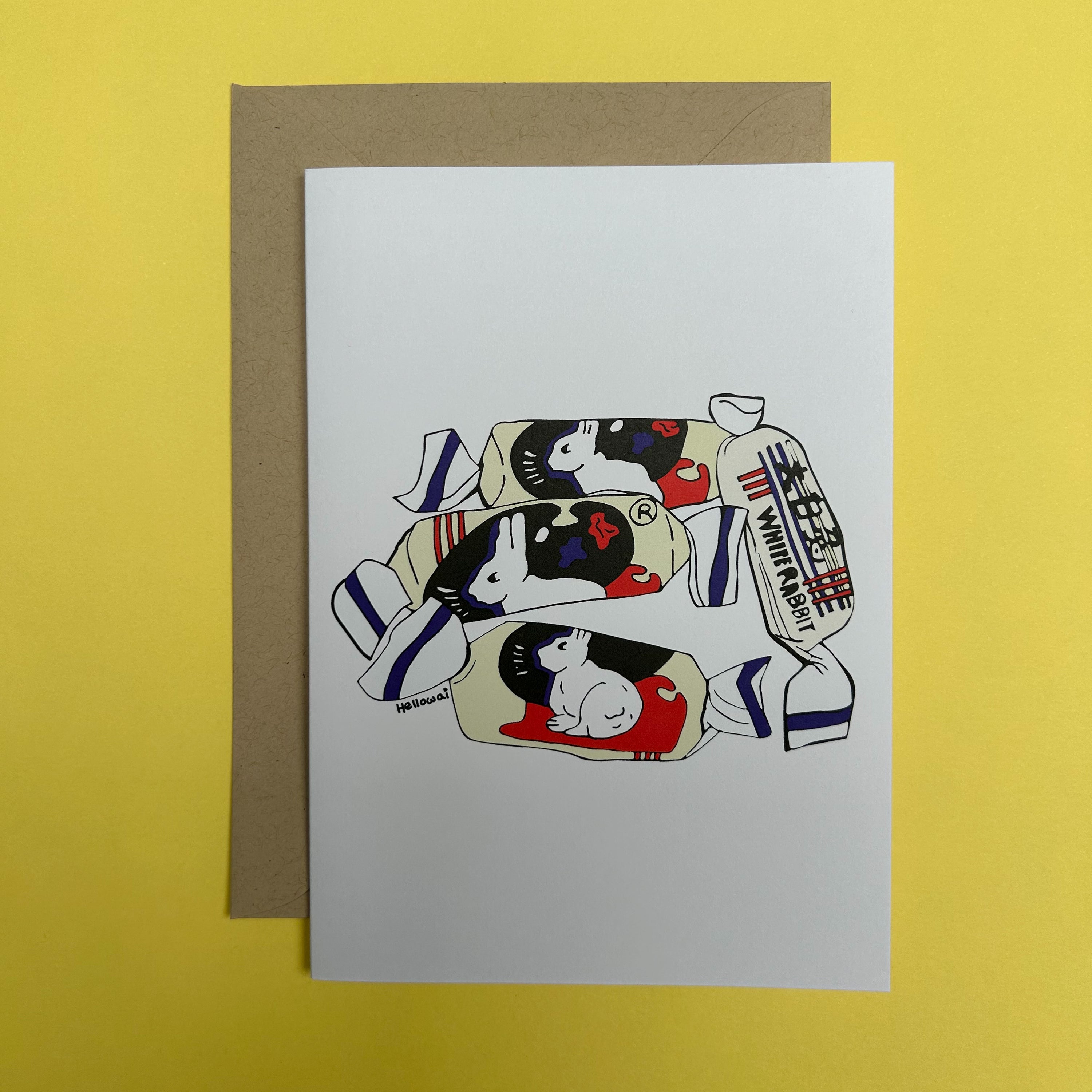 An A6 Hello Wai folded greeting card with a colour illustration of four White Rabbit Candies in their wrappers, printed on white card. The card is placed on top of a brown envelope.